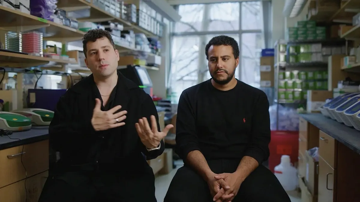 The image shows two men sitting next to each other in what appears to be a laboratory setting. The man on the left is wearing a black shirt and has his hands raised while speaking or gesturing. The man on the right has a beard and is wearing a black long-sleeved shirt, looking forward with a serious expression. In the background, shelves filled with various laboratory equipment, boxes, and supplies are visible, indicating a science or research environment