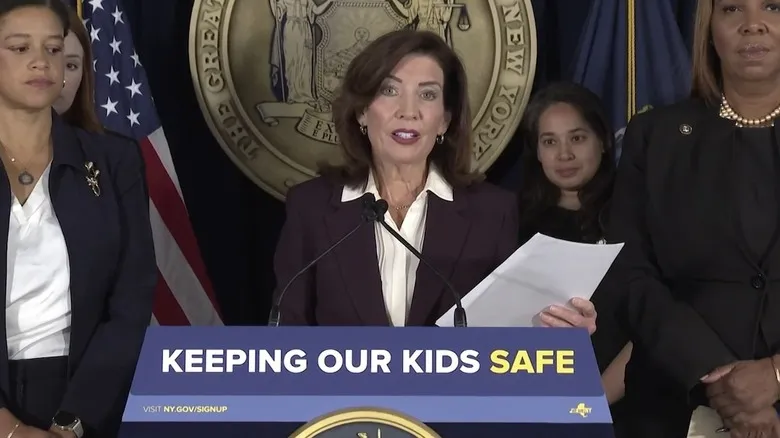حاکم نیویورک Kathy Hochul پشت یک منبر با تابلوی «حفاظت از بچه‌های ما» قرار دارد.